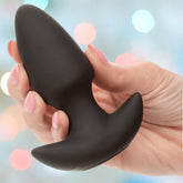 A hand with pink manicured nails holds the black CalExotics Rock Bottom Thrusting Probe against a softly blurred, colorful background.